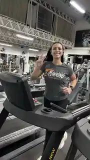 Aline ao lado de uma esteira na academia com camisa da 7RUNNING